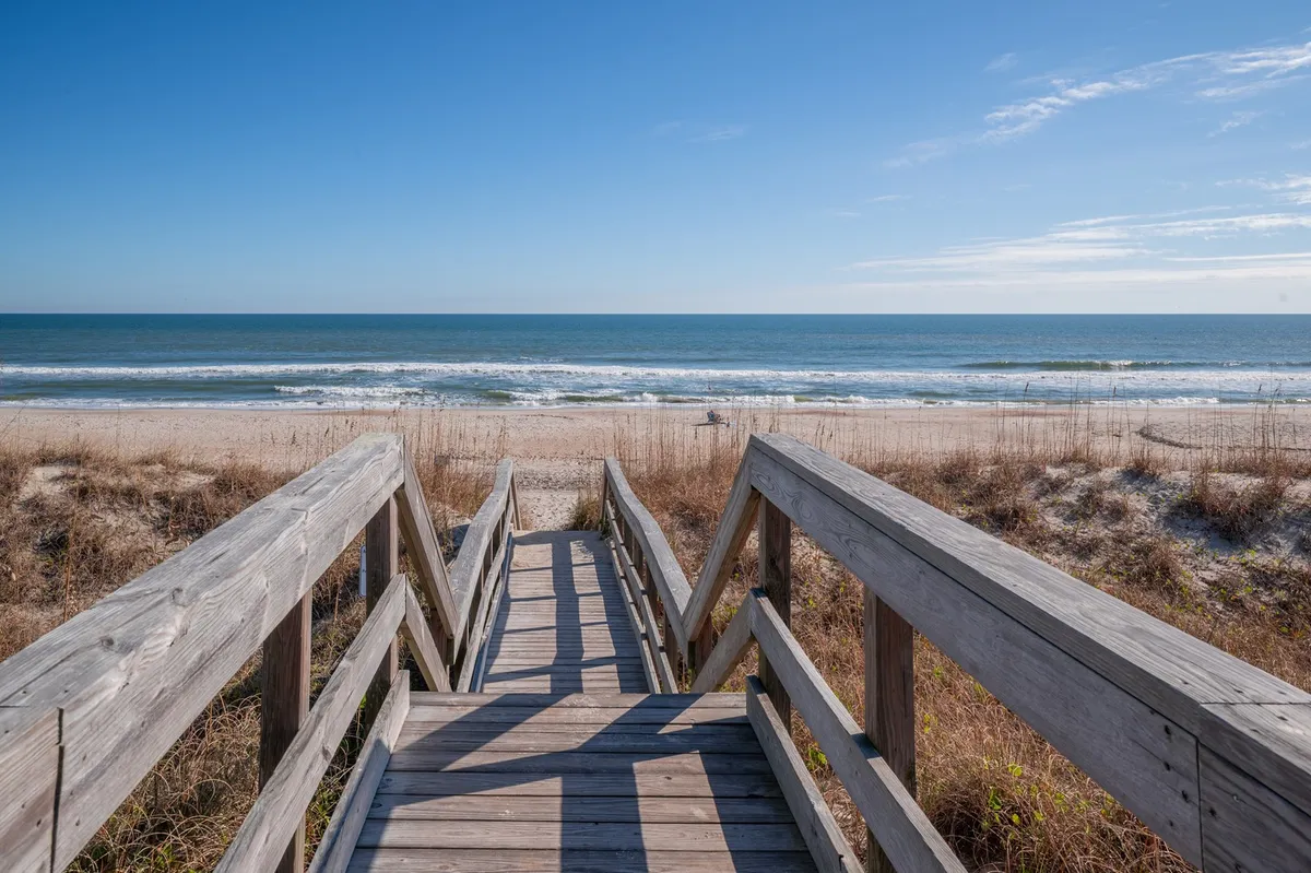 A serene view of a wooden boardwalk stretching through dune grass, leading directly to the ocean with gentle waves and a sandy shore in the distance. The sky is open and inviting, evoking a sense of anticipation for a beach adventure.