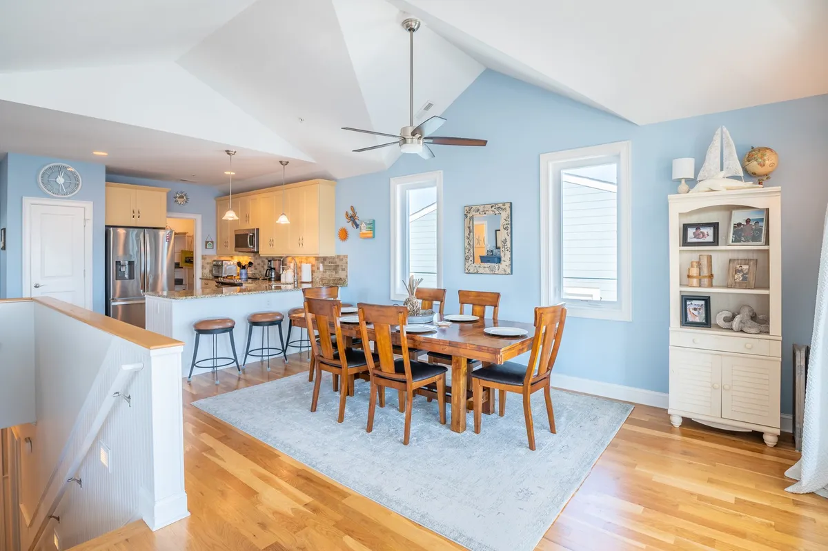 The dining area with a solid wood table sits adjacent to the kitchen, framed by large windows that fill the space with natural light. The table is set for a gathering, and the overall ambiance is inviting and cheerful, reinforcing a sense of shared meals and celebrations.