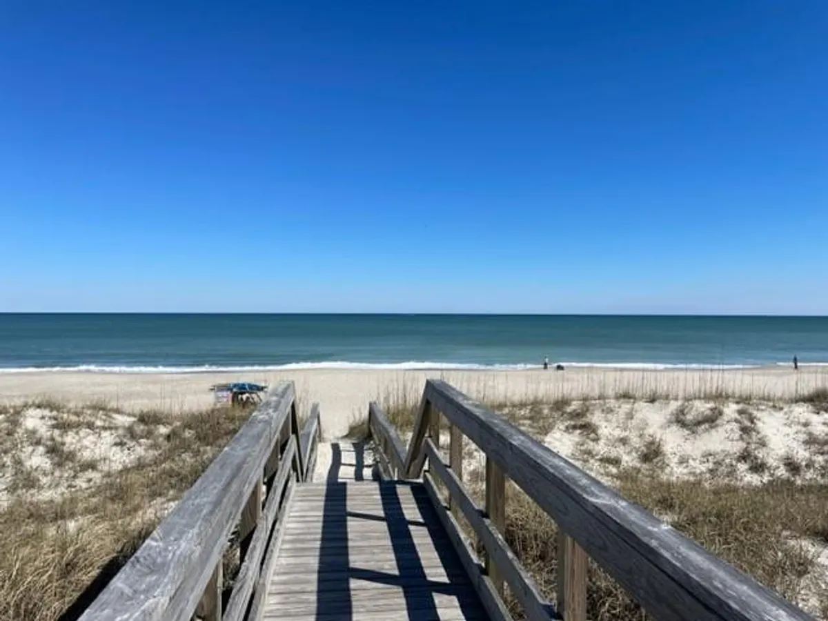A wide shot of a wooden boardwalk leading over sandy dunes to a spacious, sunlit beach. The ocean is visible beyond the dunes, and the relaxed mood suggests both family outings and peaceful morning walks.