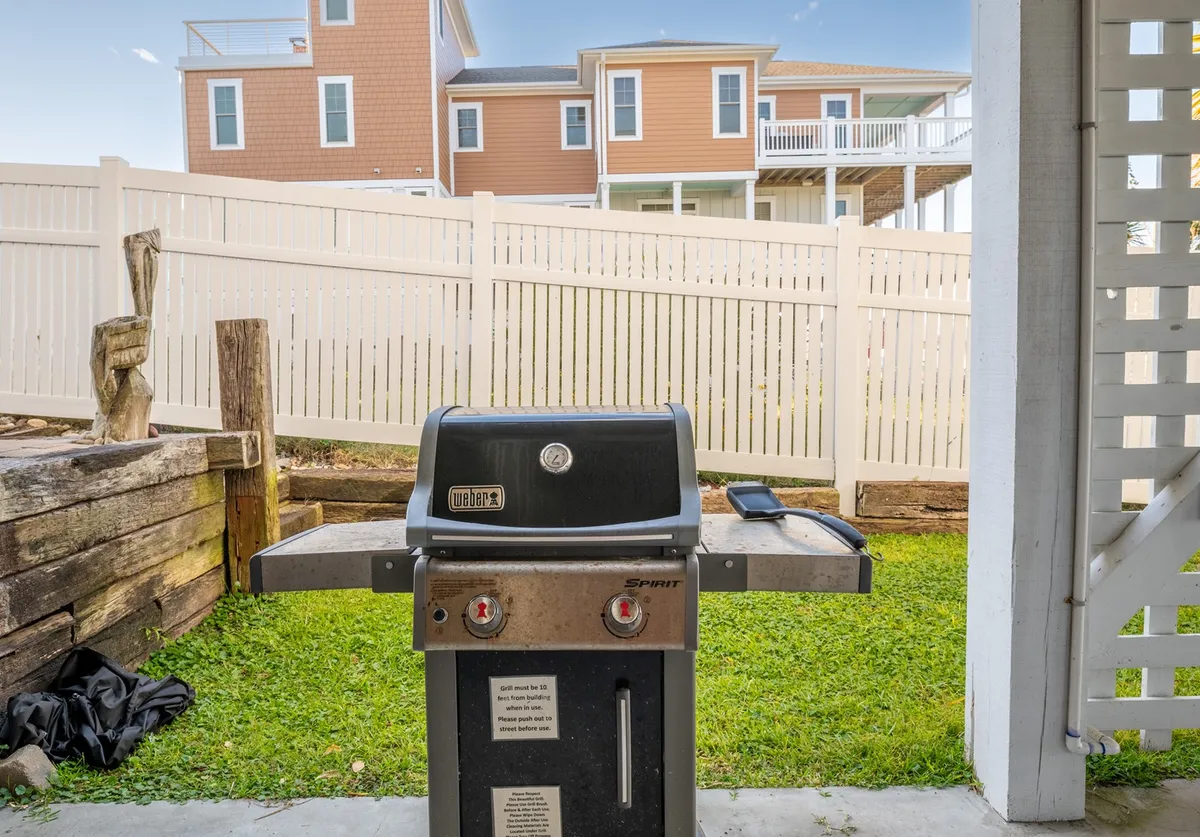 A relaxed backyard scene with a grill, green grass, and a white fence. People enjoy barbecuing and lounging outdoors, with other houses in the background suggesting a community atmosphere. The image evokes feelings of shared summer parties, celebrations, and relaxed evenings under the stars.