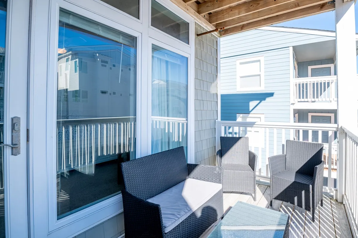 A casual outdoor seating area is shown, featuring comfortable chairs, a sliding glass door, and the blue siding of a neighboring building. The mood is relaxed and social, perfect for morning coffee, evening stargazing, or friendly chats.