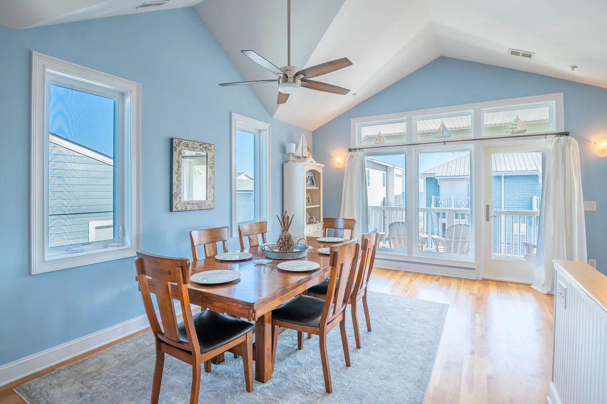 A warm, inviting dining area with a spacious table, large windows, and a ceiling fan. The setting is prepared for family meals or gatherings of friends, with natural light streaming in and the possibility of sunset views. The mood is relaxed and welcoming, evoking the sense of intimate celebrations and shared meals.