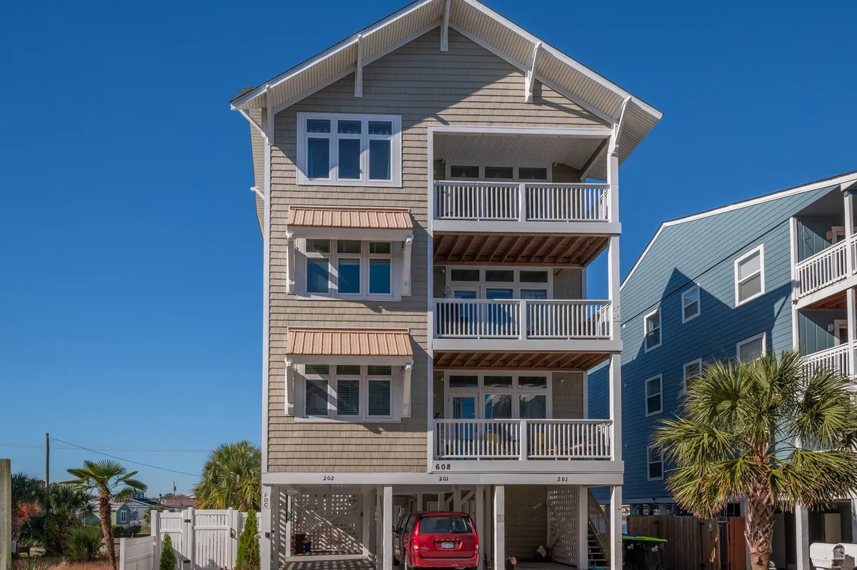 This image depicts the three-story exterior of Wood Vista, complete with balconies overlooking palm trees and a driveway with a car parked. The scene is cheerful, conveying a welcoming first impression and inviting thoughts of outdoor relaxation or family gatherings on the porches and balconies.