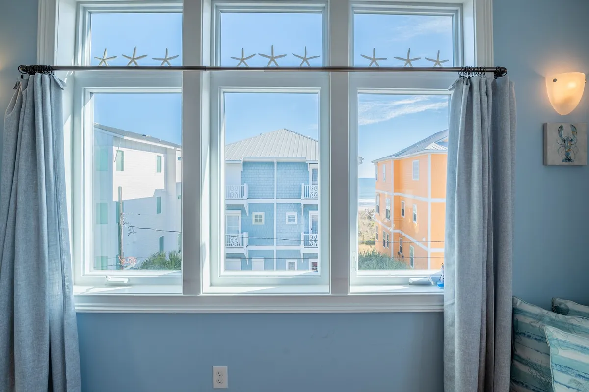 The photo captures the interior of the home, with large windows decorated with starfish, colorful coastal buildings outside, and a distant ocean view. Natural light fills the tranquil room, suggesting moments of relaxation while still connected to the outdoors.