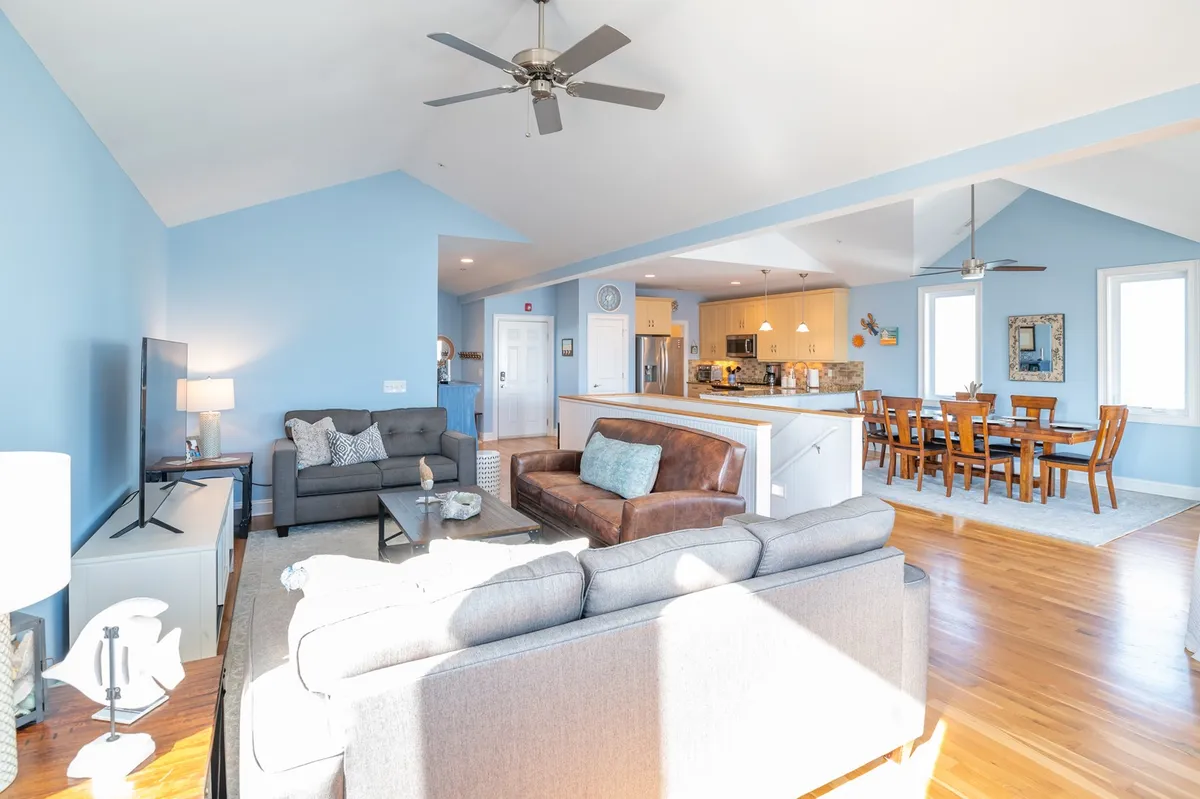 A wide view of the open-concept great room, featuring plush sofas, a spacious dining area with a solid wood table, a modern kitchen with an island, and tall windows streaming in sunlight. The arrangement promotes togetherness and allows for seamless flow between cooking, dining, and lounging.