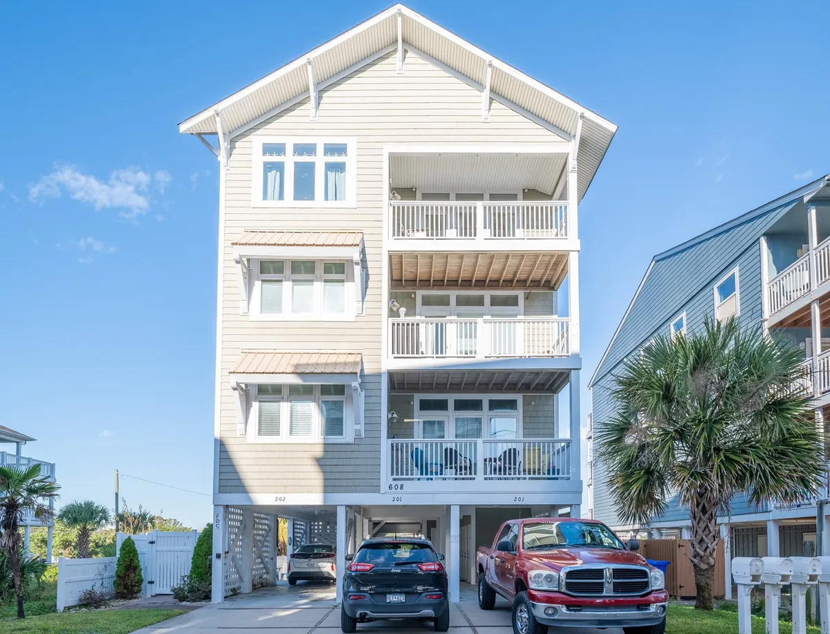 A three-story townhome building with palm trees lining the driveway and several cars parked beneath the building, emphasizing the ample, covered parking area. The balconies and inviting exterior hint at communal and resort-style living.