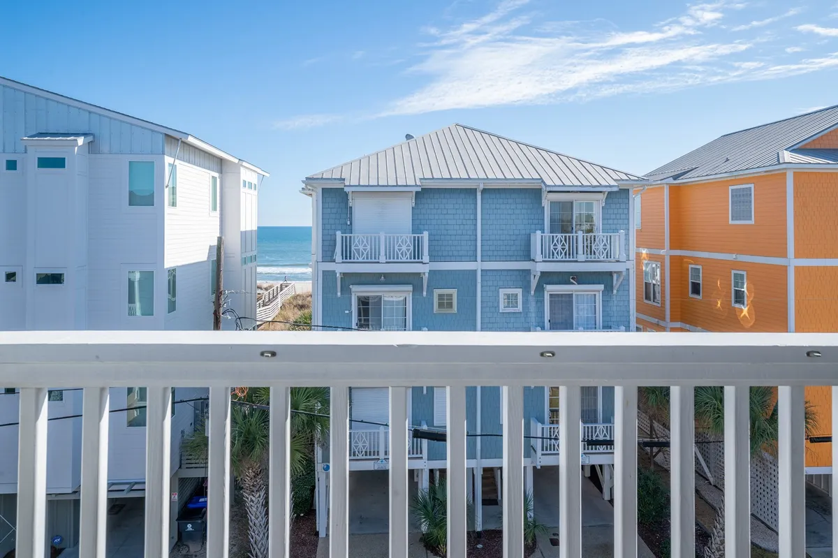 A sunny balcony scene with a clear view of the ocean, framed by a blue and orange building. The balcony railing invites guests to relax and take in the coastal scenery, perfect for enjoying the breeze or a vibrant sunset.