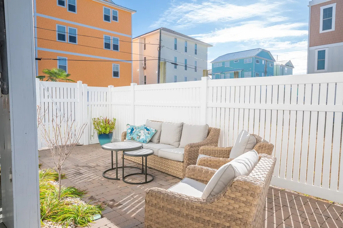 A cozy outdoor seating area on a fenced patio, complete with rattan chairs, decorative pillows, and potted plants. The space feels sunny, relaxed, and perfect for casual gatherings or morning coffee.