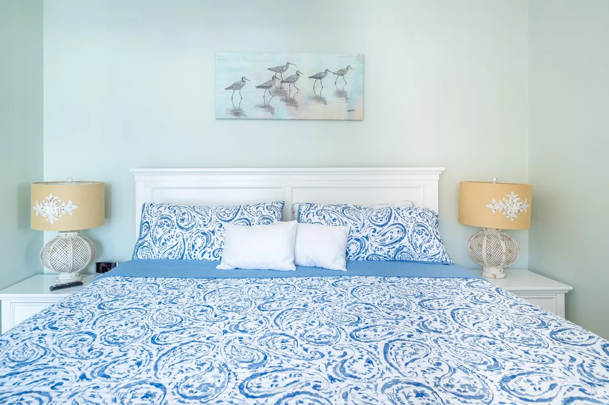 A calm and inviting queen suite with a bed featuring patterned bedding, decorative lamps, and soothing artwork. The soft colors and natural light create a restful atmosphere, perfect for unwinding after a day at the beach.