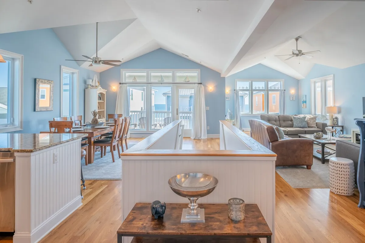 The image showcases a bright, open-concept living and dining area with a sunlit dining table set for family meals, a cozy seating area perfect for relaxation or movie nights, and large windows that let in abundant natural light. Thoughtful decorative touches create a welcoming, beachy ambiance. The scene evokes warmth and togetherness, ideal for socializing or unwinding after a day at the beach.
